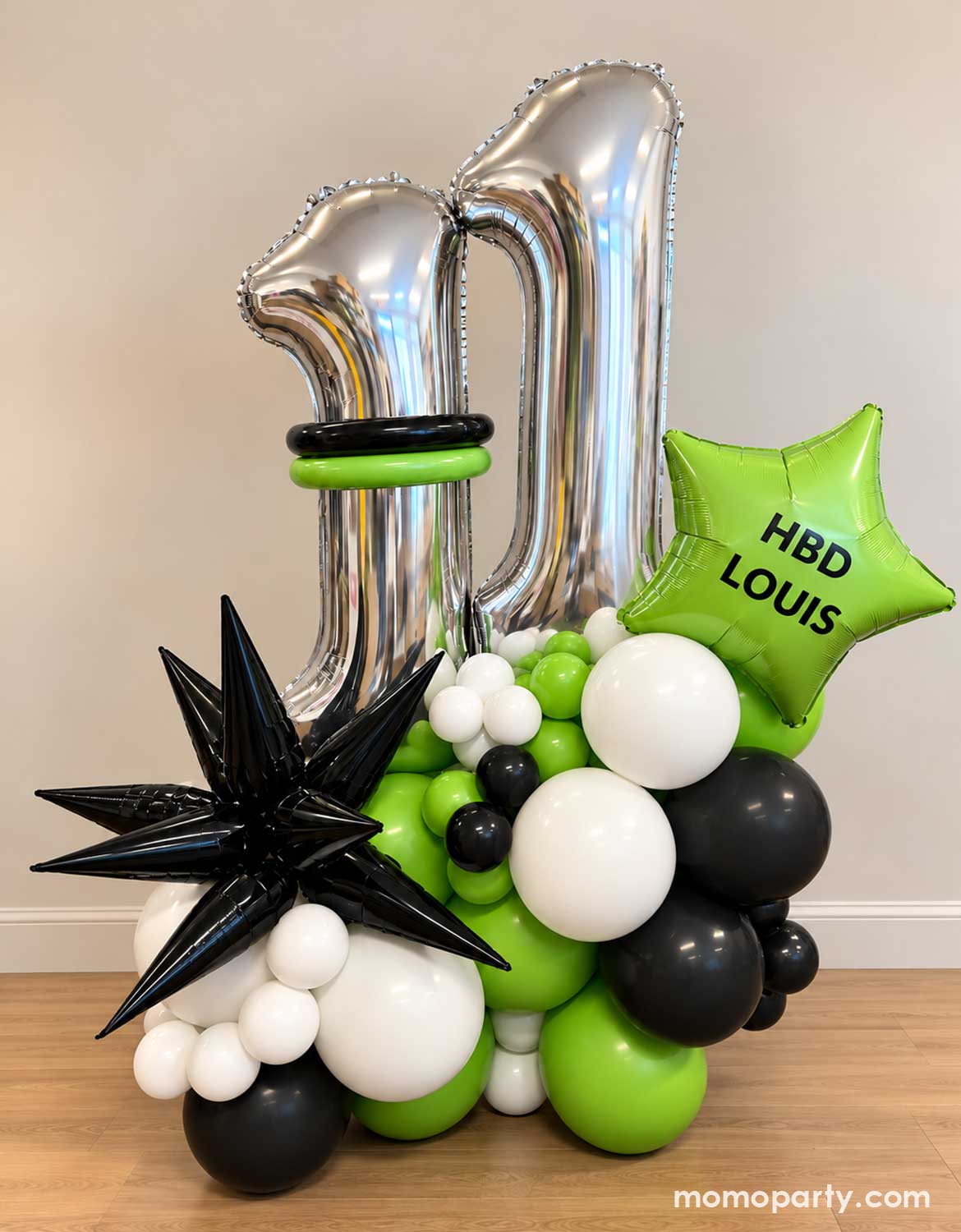 A modern and stylish birthday balloon arrangement by Momo Party for a 11th birthday featuring two large metallic silver number 11 foil balloons, a bright green star-shaped balloon with happy birthday wording a black starburst foil balloon decoration, and clusters of black, white, and lime green balloons on a wooden floor against a beige wall.