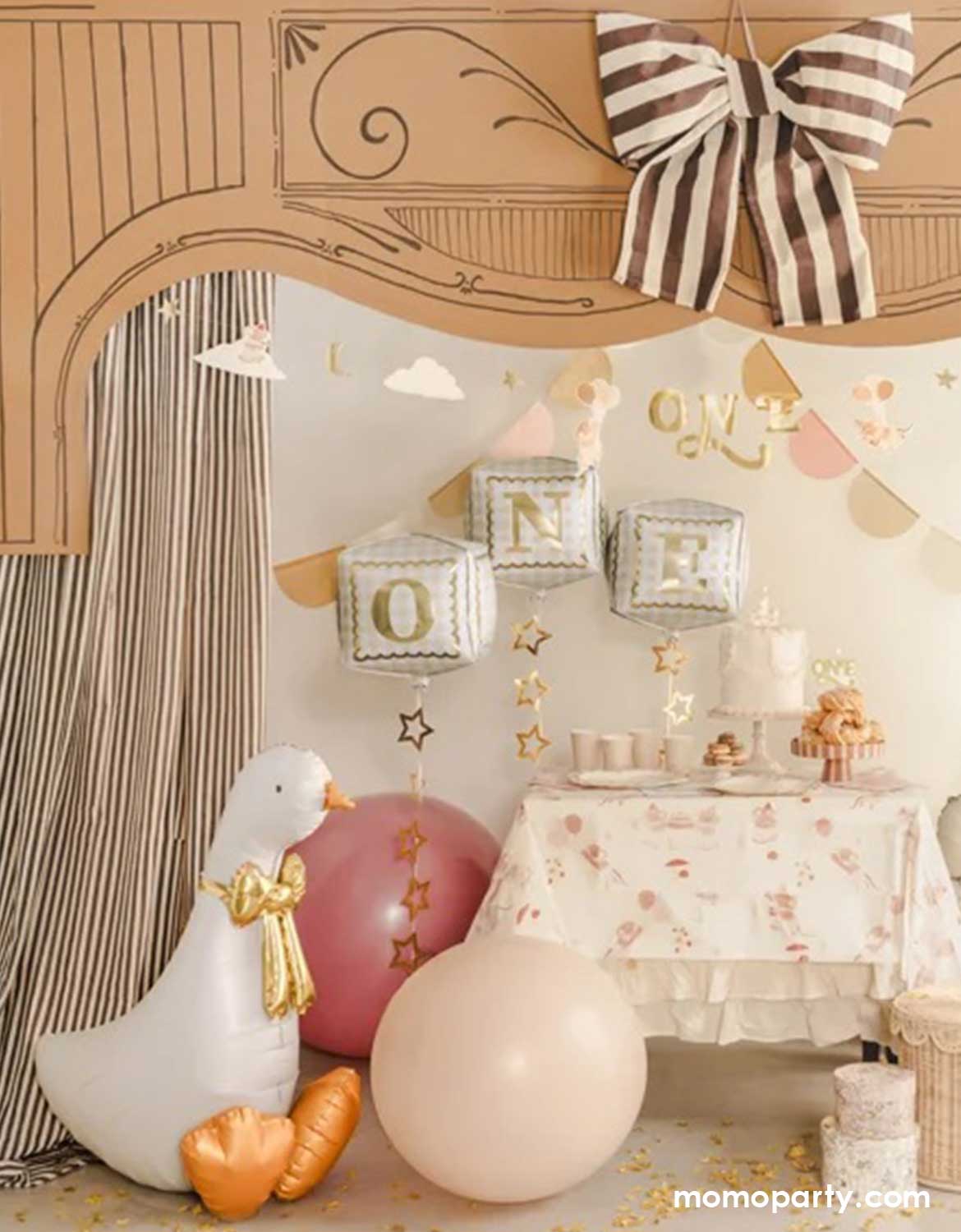 A kid's one year birthday party decoration setup featuring balloons, cake, and 'ONE' letter foil balloon decorations. Momo Party's 41" goose shaped foil balloon sitting on the ground next to two jumbo latex balloons in blush and red. With a dessert table with cakes and treats on top and party garlands hung above, this makes an adorable decorating idea for a child's first birthday party celebration.