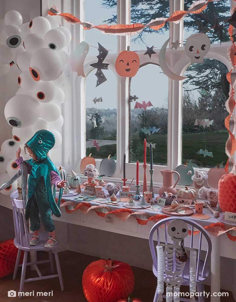 A not-so-spooky Halloween party decoration set up by Meri Meri featuring Momo Party's pastel Halloween party supplies including an eyeball balloon garland, pastel Halloween stitched streamer and pastel Halloween large garland hung on the wall. On the table was set with various pastel Halloween themed tableware including pumpkin shaped plates, napkins and party cups. In the front two kids dressed up in costume having fun at the party.