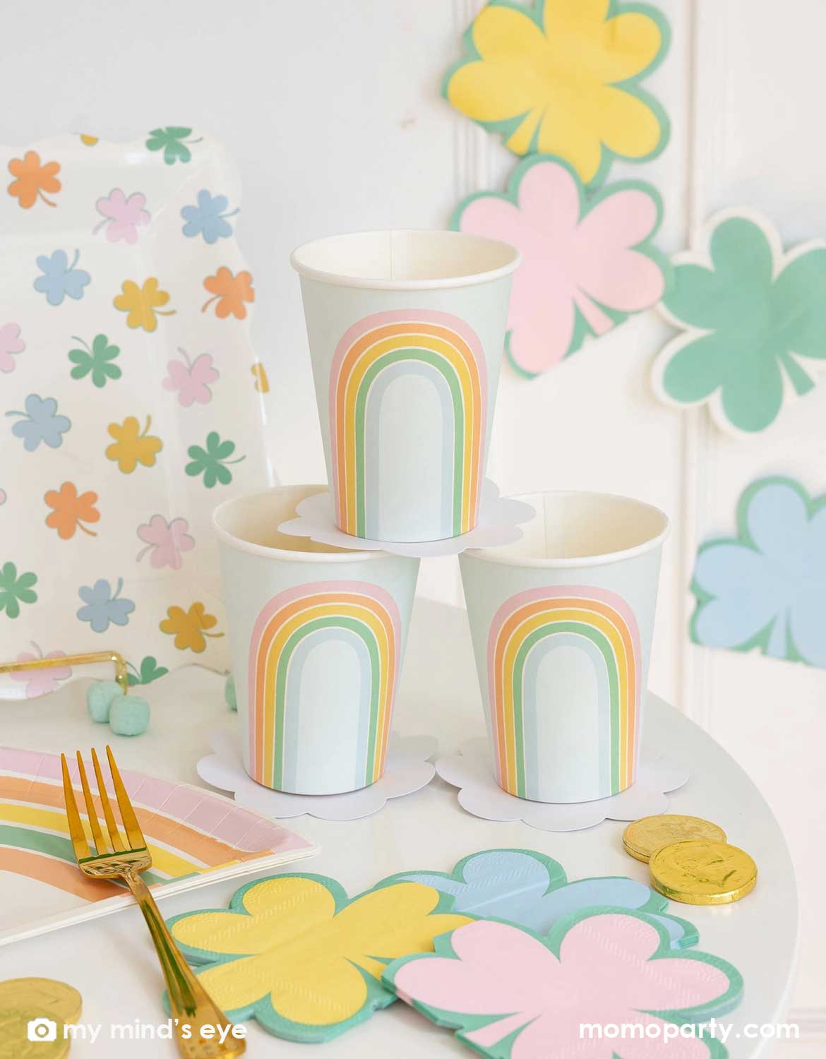A pastel themed table setting for St. Patrick'd Day celebration featuring various party supplies from Momo Party including the pastel rainbow party cups, shamrock shaped napkins, rainbow shaped paper plates, and pastel shamrock dinner plates, with chocolate coins and candies on the table, this set up is perfect for kid's St. Patrick's Day party.