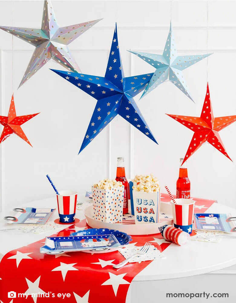 A festive Fourth of July party table decoration featuring Momo Party's Sparklers and Rockets Decorative Hanging Stars by My Mind's Eye hanging above the party table filled with stars and stripes themed patriotic party supplies including a red star pattern table runner, firework truck shaped plates, uncle sam hat party cups, rocket pop treat poppers and star patterned treat boxes with popcorn. All makes a great inspo for an unforgettable 4th of July celebration!
