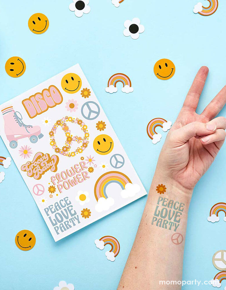 Momo Party's Peace Love Party Temporary Tattoo Sheets next to a hand with peace gesture next to it. In the light blue background there is party confetti in the shape of a smiley face, daisy and rainbow.