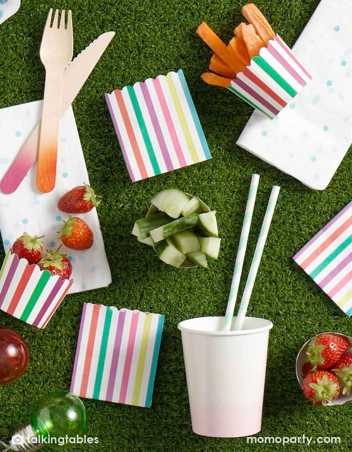 A styled picnic party setup on Momo Party's faux grass table runner with colorful striped paper cups, party plates, napkins, wooden cutlery, white straws, strawberries, cucumber wedges, and carrot fries.