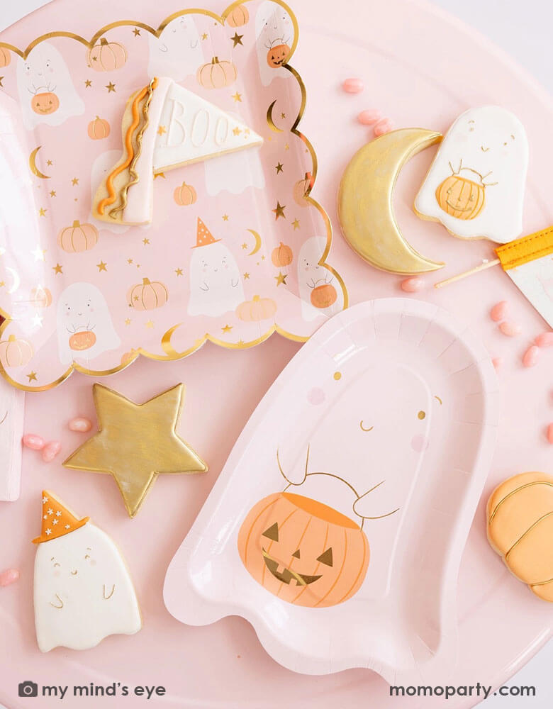A pink table filled with pink Halloween ghost themed party supplies from Momo Party including the pink ghost shaped paper plates with a jack-o-lantern, the 10" x 10" pink ghost scattered scalloped dinner plates, and a Boo felt party pennant. With matching ghost themed sugar cookies around and pink candies, it's a great inspo for a kid's friendly pink Halloween party this spooky season!