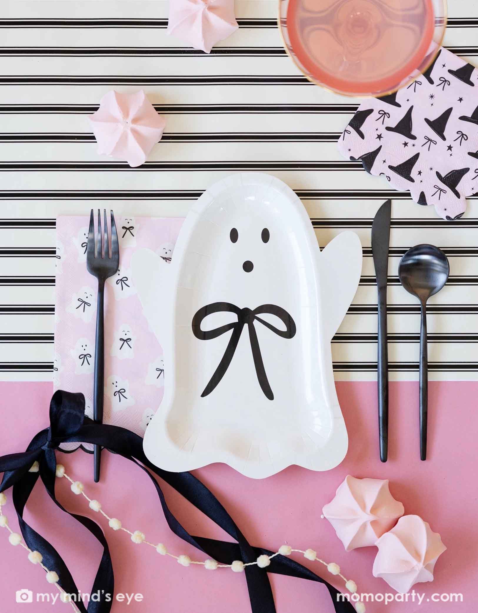 A overhead shot of a pink Halloween themed party tablescape featuring various adorable pink themed Halloween party supplies from Momo Party including the Halloween ghost plate with black bow, the pink napkin with witch hat and black bow pattern on it, black fork, knife, and spoon on a white and cream striped tablerunner with beautiful black bow around and light pink meringue desserts on the table.