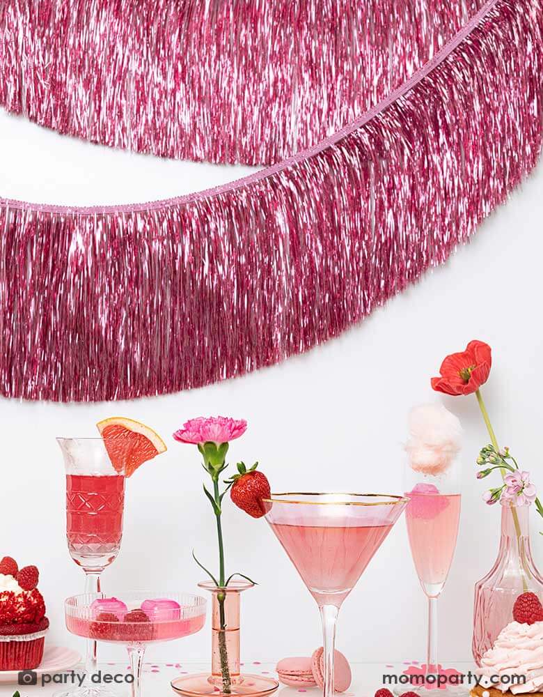 A pink themed party feature a sort of glasses with pink beverages and desserts including cupcakes, macaroons, candies and strawberries. The wall in back is decorated with Momo Party's 4.43 ft pink tinsel fringe garland by Party Deco. A perfect inspo for Valentine's Day or Galentine's Day celebrations.