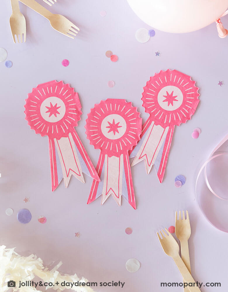 Three of Momo Party's 7.5" x 4" Show Ribbon Shaped Guest Napkins by Daydream Society on a lilac table. Around them are pink and lilac round confetti. some party balloons, party streamers and light pink ribbons.