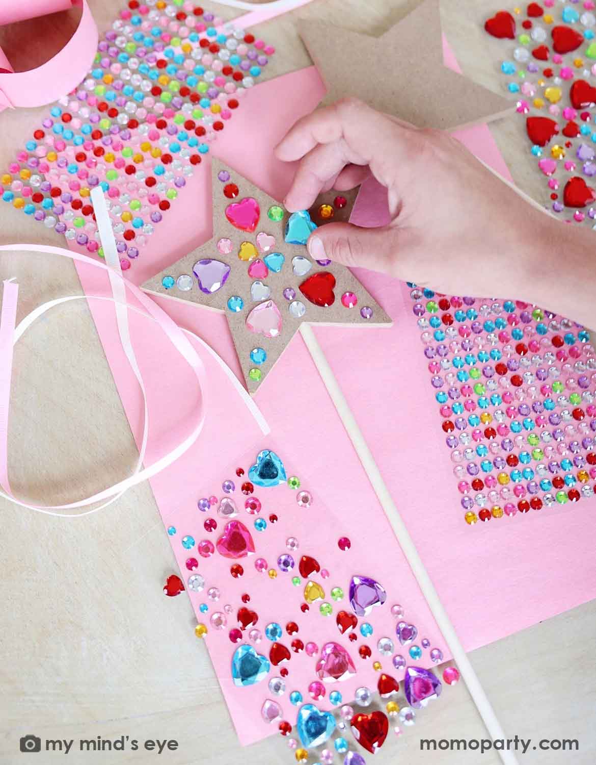 In a princess themed birthday party, a girl's hand applying colorful gemstones to Momo Party's Princess DIY Wood Wand  with pink paper and ribbons around.