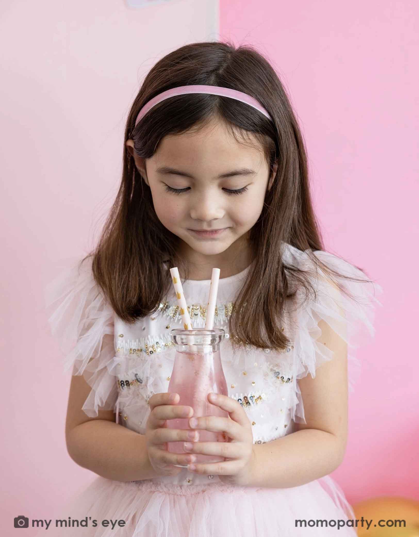 A young girl in a pale pink fairytale dress holding a pink drink with Momo Party's Princess Reusable Straws featuring gold crown pattern against a pink background.