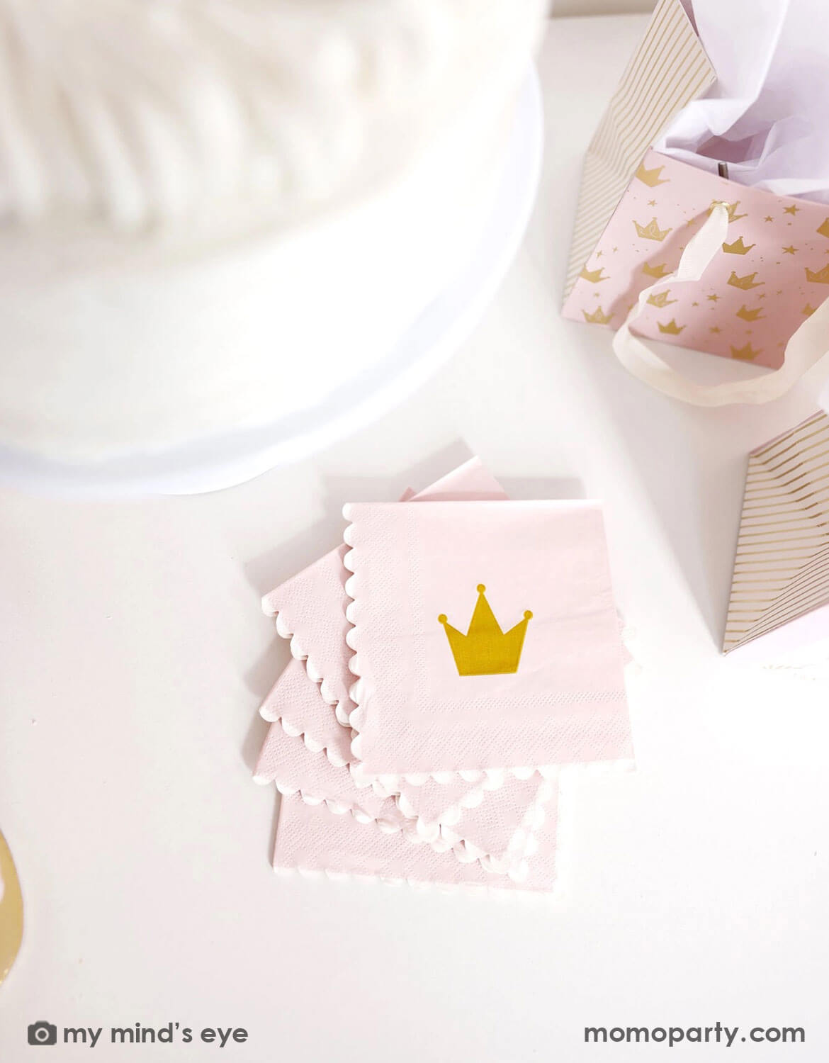 An overhead shot of kid's princess themed party table featuring Momo Party's Princess Crown Small Napkins  with gold crowns on a white table with some princess themed gift bags around.