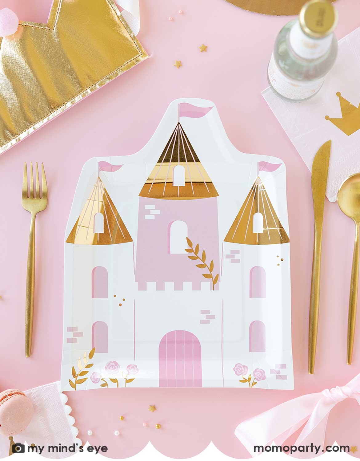 An overhead shot of a kid's princess birthday party table featuring Momo Party's princess castle shaped plate, crown napkins, decorative crowns and gold utensils around. With decorative elements and treats around on a pink tablecloth, this is a great inspiration for parents who are throwing a fairytale themed party for their kids.