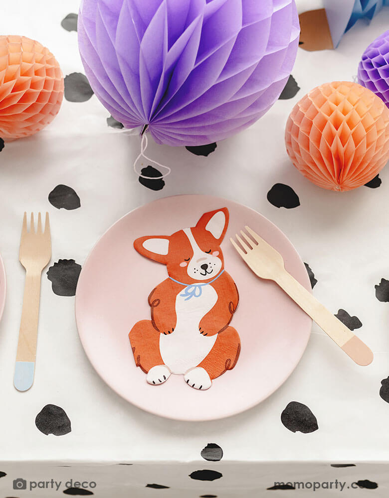 A close up of a dog themed party tablescape featuring a Corgi shaped napkin on a pink round plate on a Dalmatian dog printed tablecloth. Around the plate there are some honeycomb ball decorations in soft pastel colors, making this an adorable inspiration for a cute dog puppy themed celebration.
