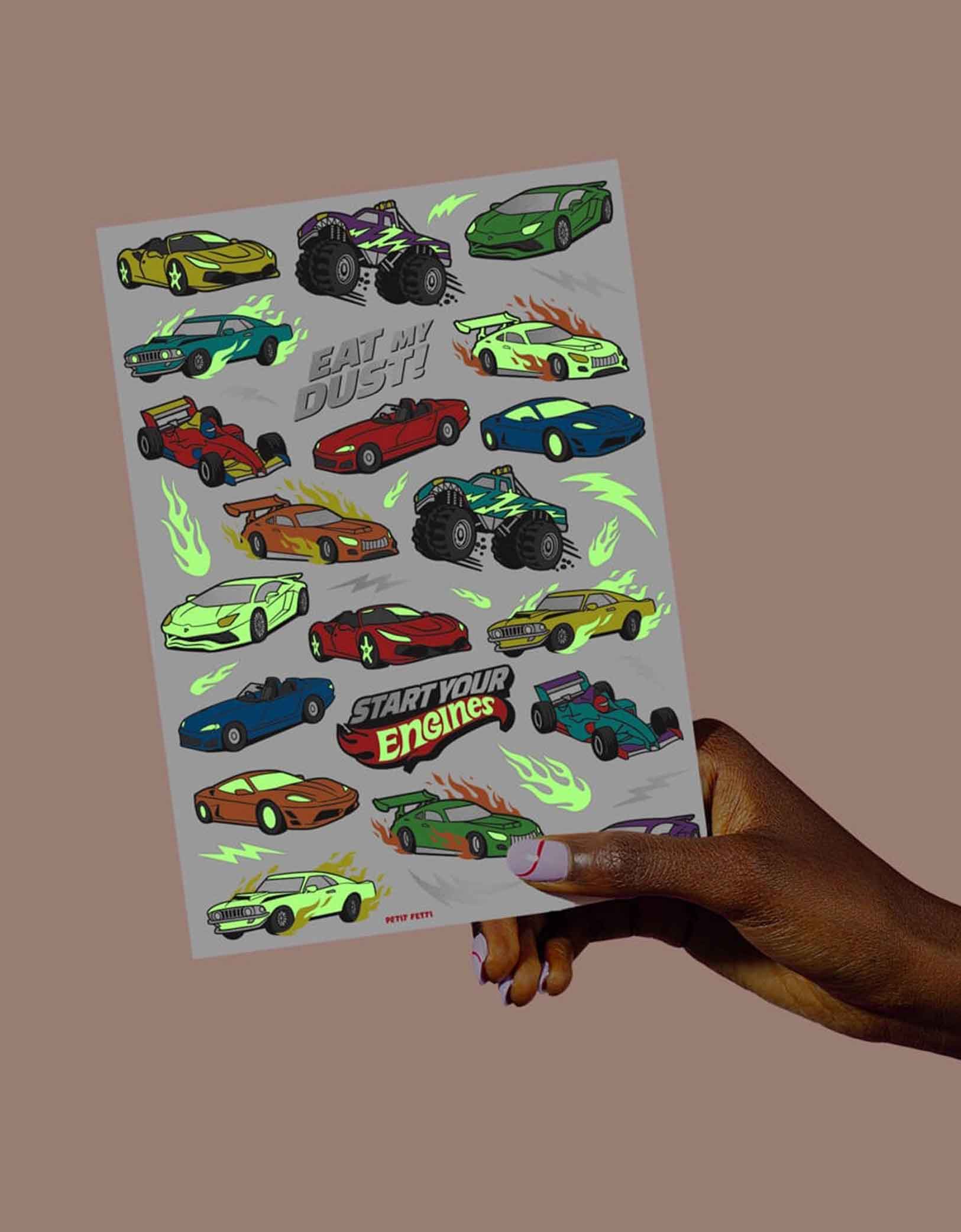 A hand is holding a sheet of colorful race car themed temporary tattoos from Momo Party with texts "Eat my dust" & "Start Your Engines" glow in the dark in a dark background.