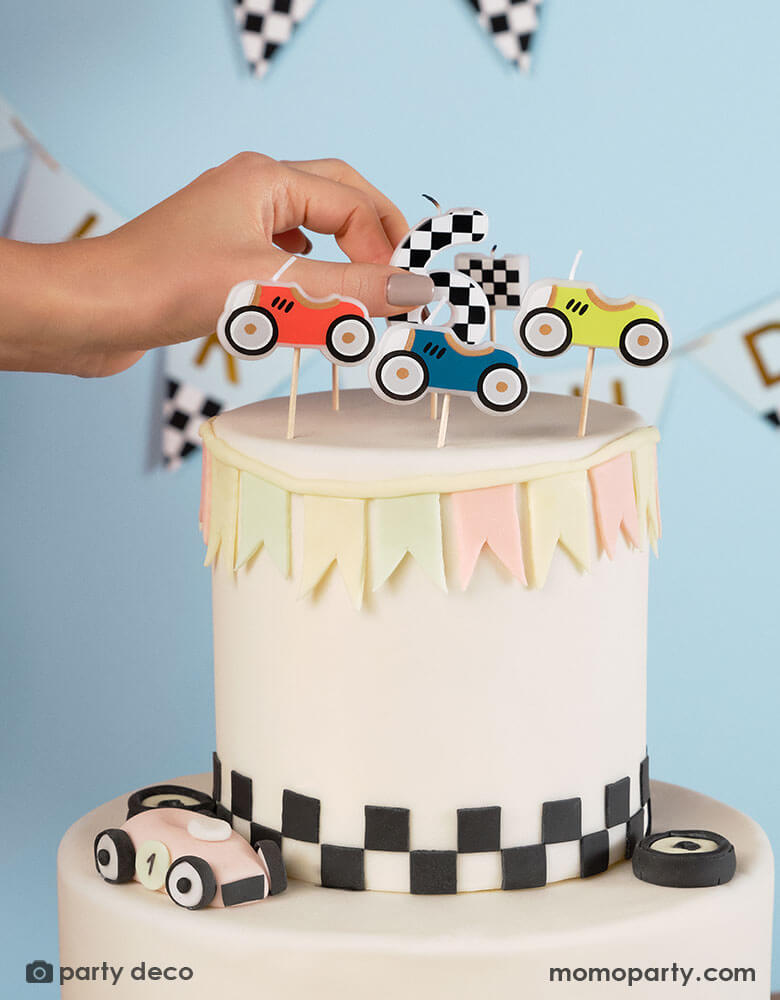 A race car themed birthday cake decorated with checkered pattern at the bottom topped with Momo Party's race car candles, a hand put a checkered number 6 candle. Makes it a great inspiration for kid's race car birthday party idea.