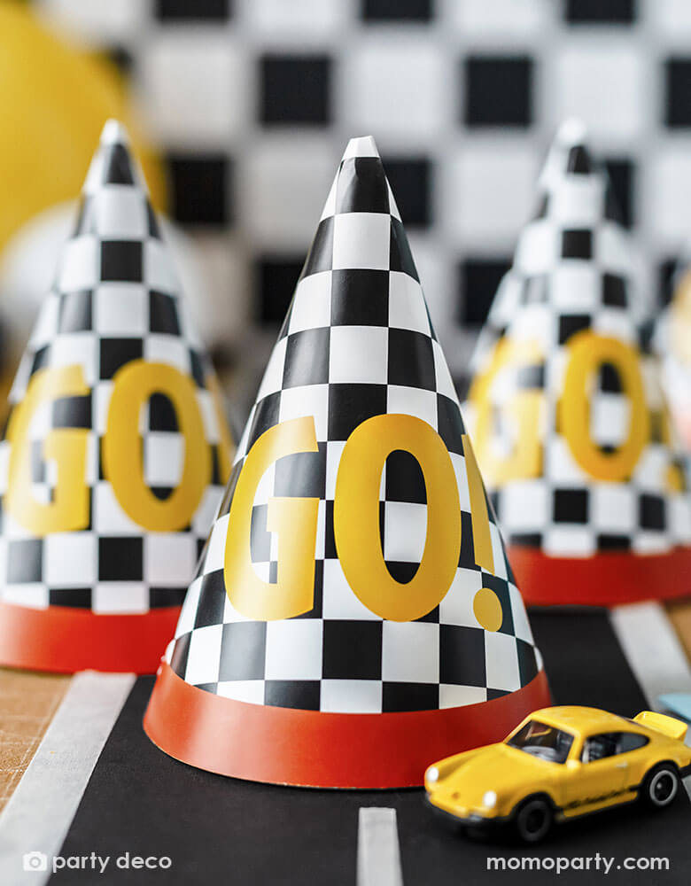 A close up shot of Momo Party's 4.3" x 6.3" race car checkered flag GO! party hats by Party Deco on party table decorated with a race car track sticker . Featuring the classic race car flag checkers with a red rim, each hat has a GO! written on it, making these a fun and festive addition to kid's race car themed celebration.