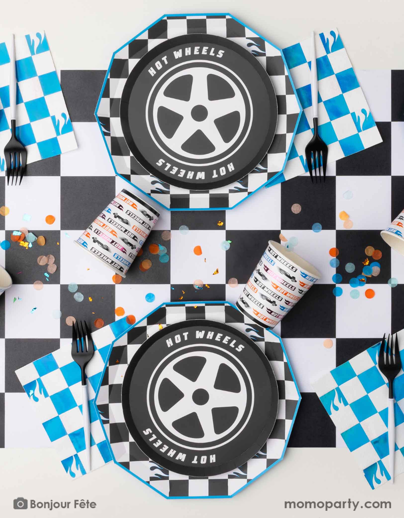 An overhead shot of a Hot Wheels-themed party table setting with tire shaped plates, checkered dinner plates, blue checkered napkins, and Hot Wheels party cups in multiple colors on a checkered table runner from Momo Party. With festive confetti spread out, this is a perfect inspiration for kid's race car themed birthday party.