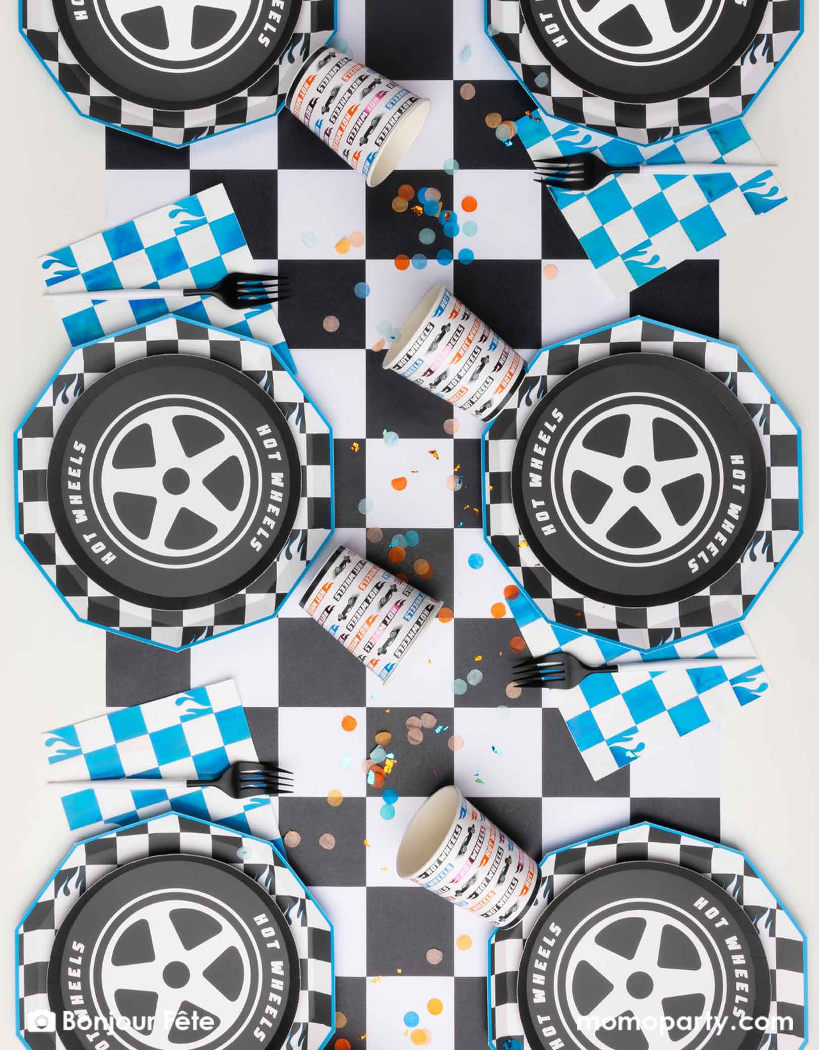 An overhead shot of a Hot Wheels-themed party table setting with tire shaped plates, checkered dinner plates, blue checkered napkins, and Hot Wheels party cups in multiple colors on a checkered table runner from Momo Party. With festive confetti spread out, this is a perfect inspiration for kid's race car themed birthday party.