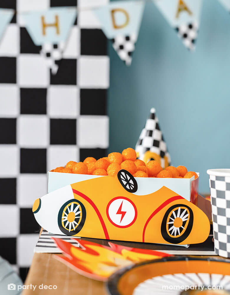 A modern kid's race car themed party table features Momo Party's race car snack box in yellow, filled with cheese ball snacks. On the table there are checkered pattern party cups, party hats and wheel shaped plates by Party Deco on a race track table runner. In the back there's a checkered backdrop decorated with a trophy "Happy Birthday" party banner in light blue, making this a perfect inspo for kid's race car themed parties.
