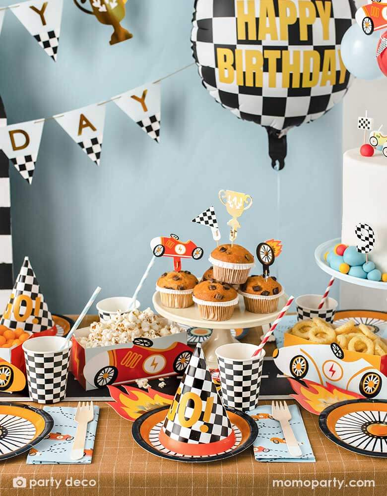 A kid's race car themed party table filled with race car themed party supplies from Momo Party including wheel shaped plates, checkered flag GO! party hats, blue race car large napkin, and checkered party cups. On the table there are muffins topped with race car themed cupcake toppers and vintage race car shaped snack holder filled with popcorn and onion ring chips. Above the table, there's a race car flag themed birthday banner and checkered happy birthday foil balloon.