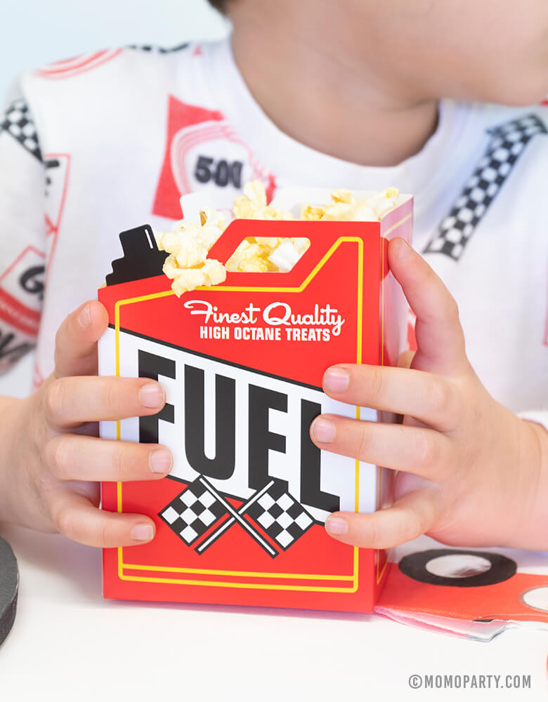 A boy dressed in a race car themed sweat shirt holding Momo Party's fuel snack box filled with popcorn in a race car themed party.