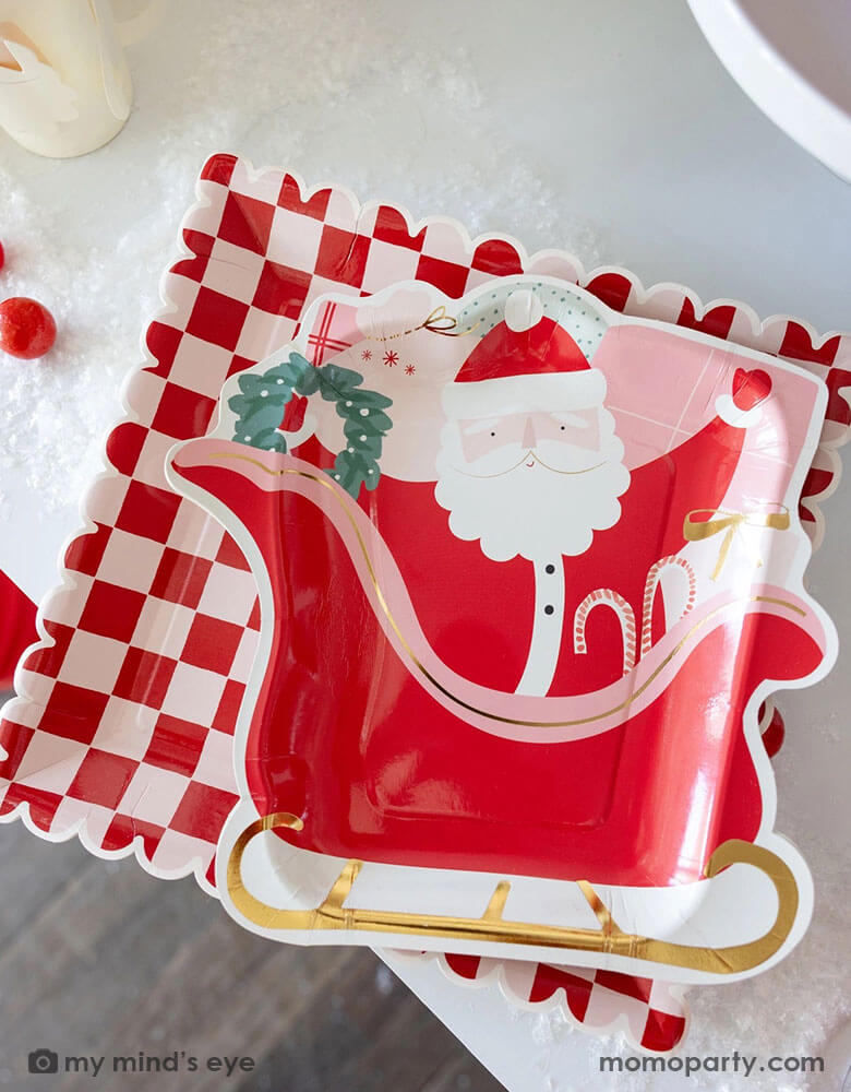 A table shot of Momo Party's 10" Pink and Red Checked Paper Plates and the Santa on a sleigh shaped plate by My Mind's Eye. These playful Santa inspired plates add a touch of fun to any table setting. Get ready to serve up some serious style with these quirky and eye-catching plates this Holiday season!