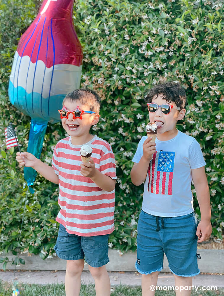 2 boys holding ice cream and United States Flag in the Backyard 4th of July Celebration Party. One boy wearing star shaped sunglasses, red and white striped T shirt and jeans short, another boy wearing a Zara T-Shirts With Sequinned Flag, There is a Anagram Balloons 40" Red White Blue Popsicle Foil Balloon on the back, So fun for 4th of July celebration