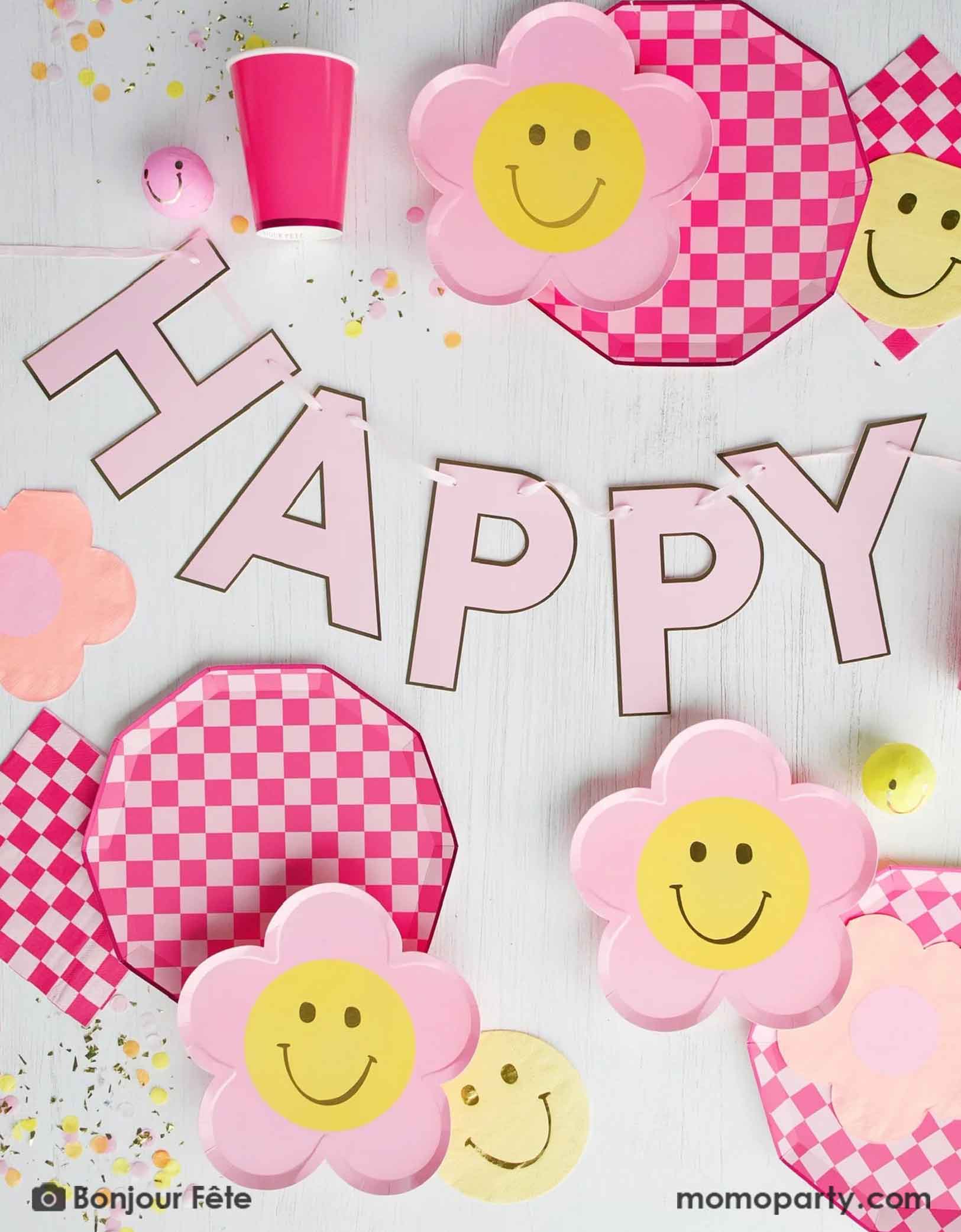 A retro inspired table setting for a kid's party featuring red and pink checkered paper plates with flower-shaped smiley faces, with a light pink "Happy Birthday" banner in the middle with confetti around from Momo Party on a light background.