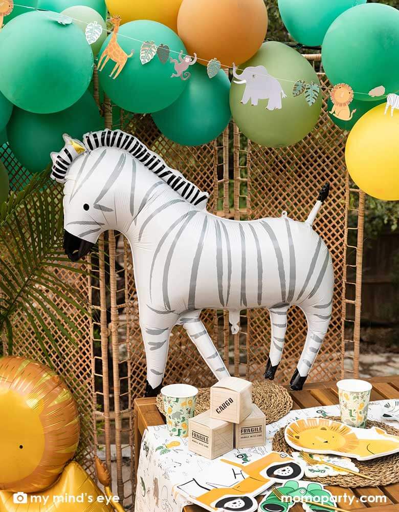 A festive kid's safari themed birthday party set up feature Momo Party's 24" zebra shaped foil mylar balloon by My Mind's Eye. Above it is a safari themed balloon garland in green, orange and yellow adorned with safari animal birthday banner. On the table, there are multiple safari themed tableware including a lion shaped plate, a Jeep car shaped plate, a binocular shaped dinner napkin and safari animal party cups on a safari themed paper table runner.