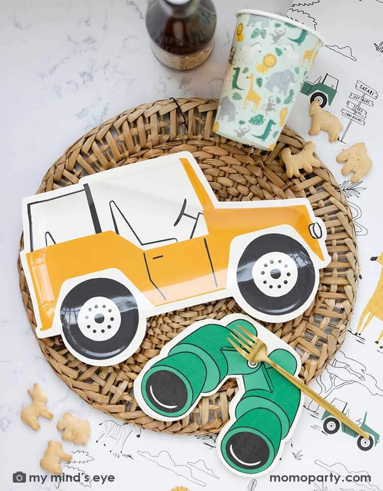 A safari adventure themed party table featuring Momo Party's mustard Jeep shaped dinner plate laid on top of a round bamboo weaved round tablemat which is placed on My Mind's Eye's safari adventure paper table runner featuring safari animals and safari landscape. Around the plate are a mint party cups with lots of safari animals and Momo Party's green binoculars shaped dinner napkins. With animal crackers on the table, makes this tablescape a great inspiration for kid's safari themed party.