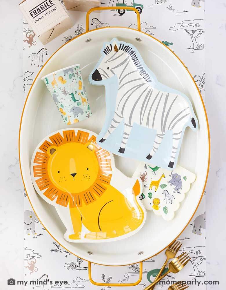 A safari adventure themed party table with an oval mustard edged plate with Momo Party's lion shaped plate and the zebra shaped plate, along with My Mind's Eye's safari party cups and small napkins, all laid on top of My Mind's Eye's safari adventure paper table runner featuring safari animals and safari landscape. Around the plate With My Mind's Eye's cargo favor boxes and gold utensils on the table, makes this tablescape a great inspiration for kid's safari themed party.