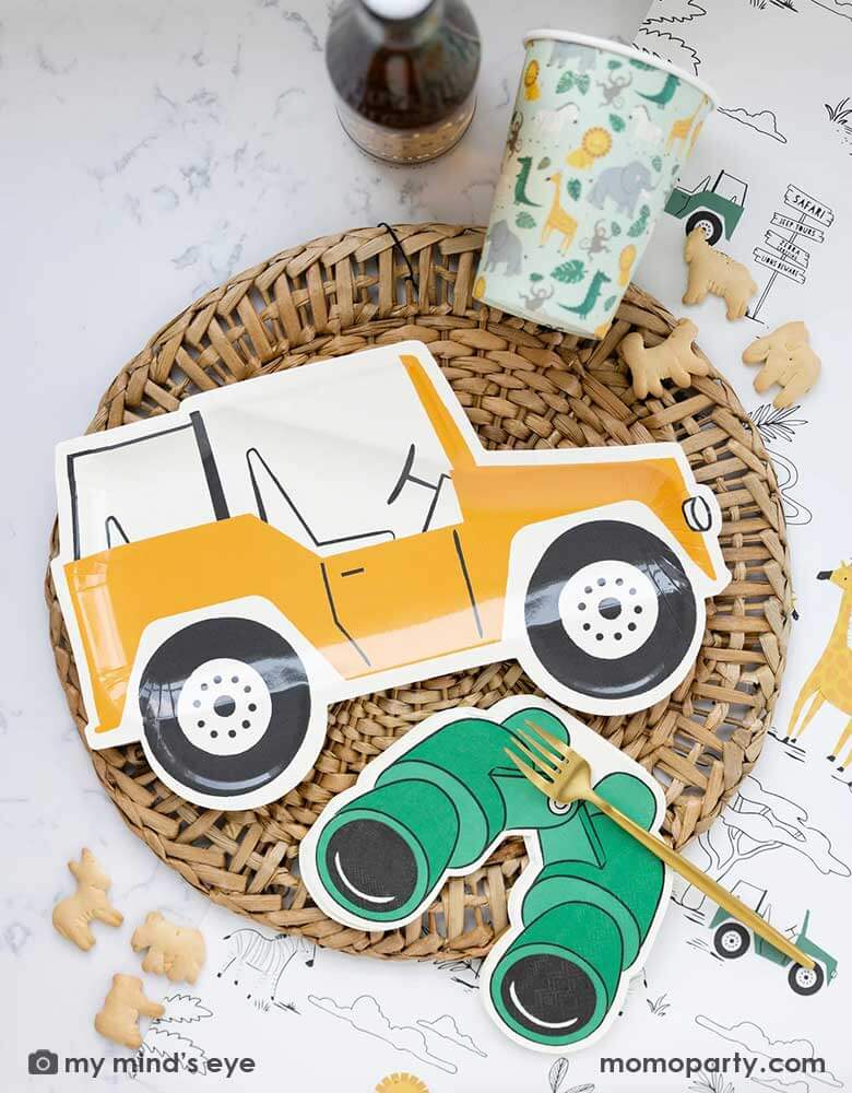 A safari adventure themed party table featuring Momo Party's mustard Jeep shaped dinner plate laid on top of a round bamboo weaved round tablemat which is placed on My Mind's Eye's safari adventure paper table runner featuring safari animals and safari landscape. Around the plate are a mint party cups with lots of safari animals and Momo Party's green binoculars shaped dinner napkins. With animal crackers on the table, makes this tablescape a great inspiration for kid's safari themed party.