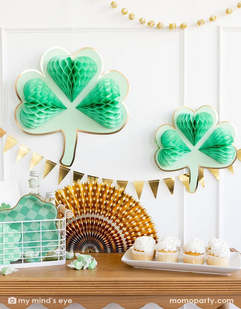 A corner of a room decorated with two of Momo Party's shamrock honeycomb decorations and a gold foil flag banner hung on the wall. On the cabinet counter, there's a gold foil paper party fan, a few cupcakes and a basket filled with Momo Party's St. Patrick's Day party tableware including scalloped paper plates, lucky paper plate set, checkered shamrock shaped napkins and plates next to some green taffy candies, makes this a perfect info for a festive St. Patrick's Day celebration.