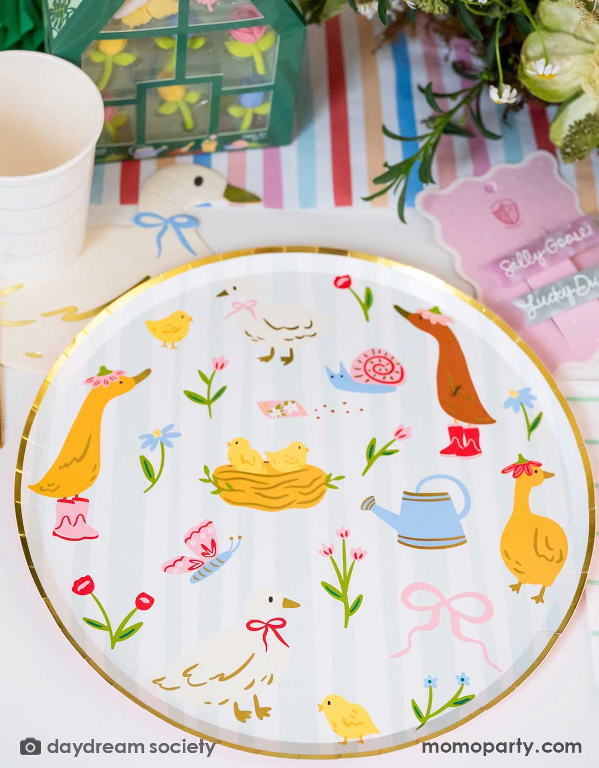 A closeup of Momo Party's 10" silly goose paper plate featuring adorable spring little animal illustrations in an Easter table setting with colorful flowers, tulip honeycomb decorations, goose themed party plates, napkins, party cups, pink cutlery and multiple kid's Easter small gifts with bunny designs on a colorful striped runner.