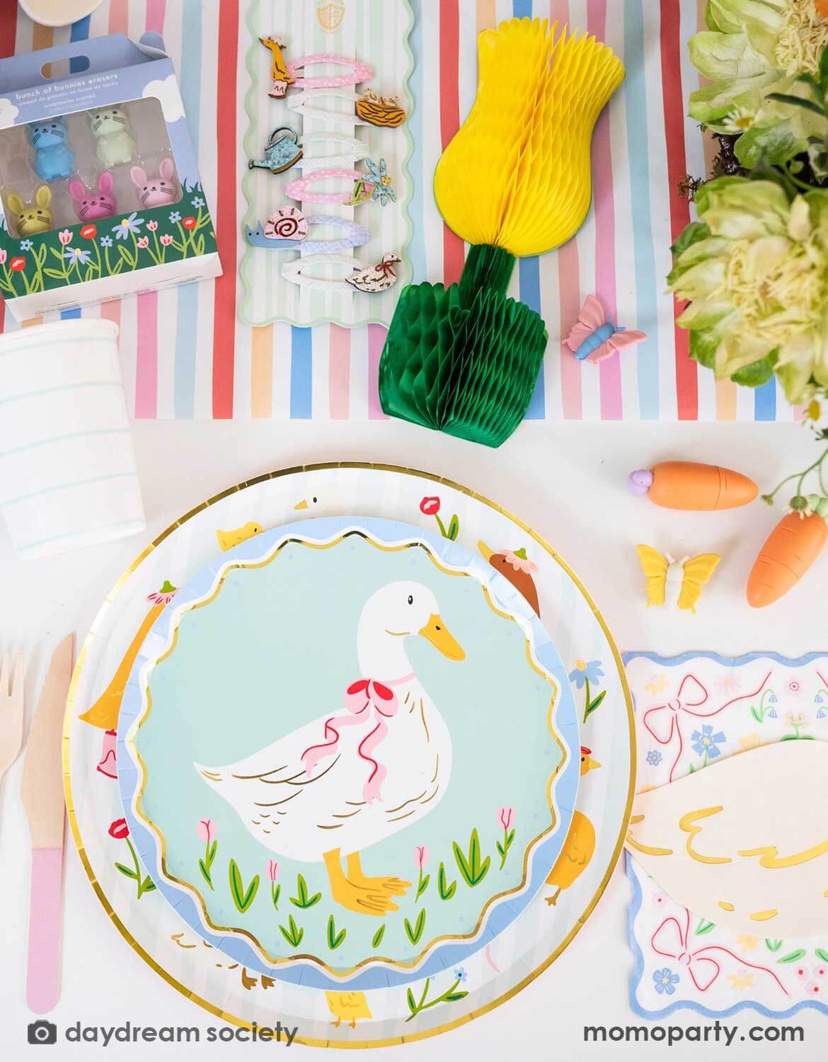 An overhead shot of a children's Easter table setting with colorful flowers, tulip honeycomb decorations, goose themed party plates, napkins, party cups, pink cutlery and multiple kid's Easter small gifts with bunny designs on a colorful striped runner. A perfect idea for an Easter celebration, a baby shower, kid's Silly Goose themed birthday party or a spring gathering.