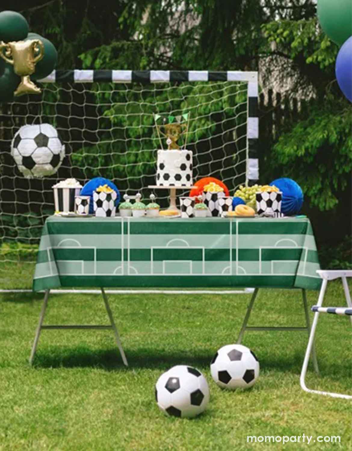 A kid's soccer-themed party setup on a grassy lawn, featuring a table covered with a green and white soccer field print tablecloth, decorated with soccer balls, a cake, popcorn, cups, and a small trophy. A black-and-white soccer goal net and balloons are in the background.