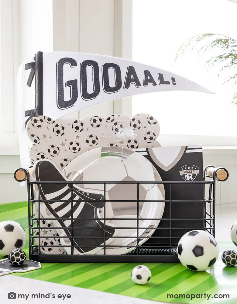 A festive occasions bin for a fun soccer party featuring Momo Party's silver soccer ball shaped plates, soccer cleats shaped napkins, black soccer jersey shaped treat bags and a black and white GOAL felt party pennant by My Mind's Eye. With a green striped paper runner underneath and some soccer ball shaped candies around the table, this makes a perfect inspo for kid's soccer themed birthday party or championship celebration.