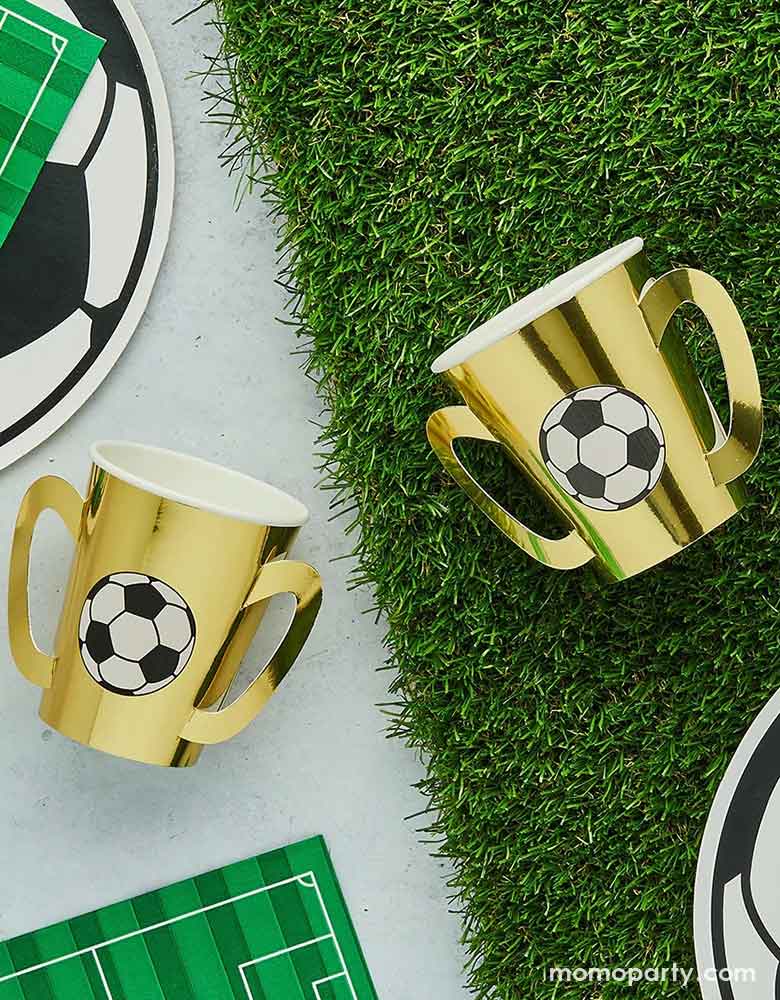 A soccer theme party table featuring soccer themed party supplies from Momo Party including gold trophy party cups with soccer ball design on them on a faux grass runner, surrounded by soccer ball shaped plates, soccer field shaped napkins, a perfect inspo for soccer themed gathering, including kid's soccer birthday parties and viewing parties.