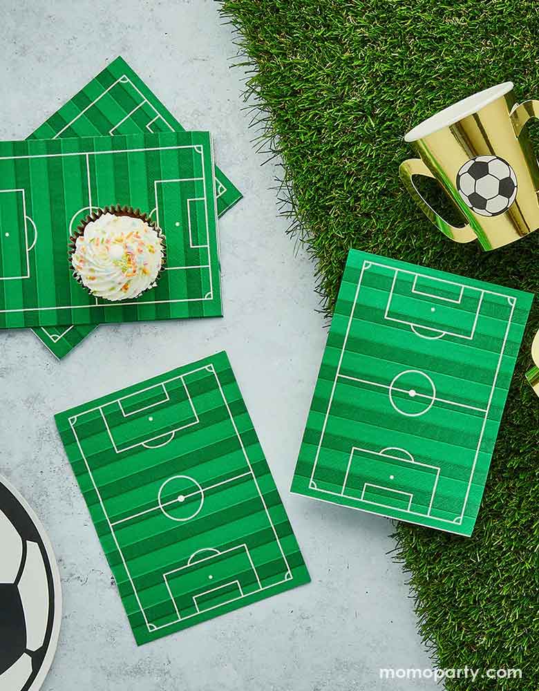 A soccer theme party table featuring soccer themed party supplies from Momo Party including soccer field dinner napkins on a faux grass table runner, surrounded by soccer ball shaped plates and gold trophy shaped party cups with soccer ball design on them, a perfect inspo for soccer themed gathering, including kid's soccer birthday parties and viewing parties.