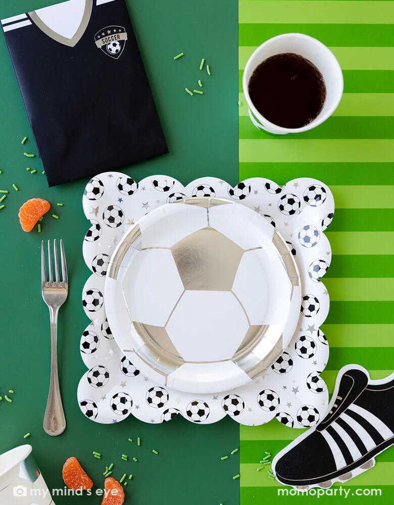 A fun soccer themed party table featuring Momo Party's soccer party collection including silver soccer shaped plates and party cups, cleat shaped napkins, soccer jersey shaped treat bags and soccer table runner by My Mind's Eye. With some snacks, green sprinkles around, this makes a great inspo for a fun kid's soccer themed birthday party or a championship celebration!