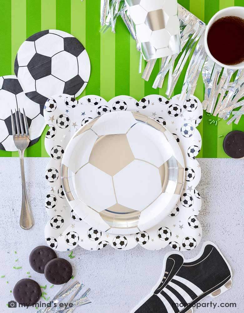 A fun soccer themed party table featuring Momo Party's soccer party collection including silver soccer shaped plates and party cups, soccer shaped napkins, cleat shaped napkins and soccer table runner by My Mind's Eye. With some snacks, silver streamers, green confetti and silver utensil around, this makes a great inspo for a fun kid's soccer themed birthday party or a championship celebration! 