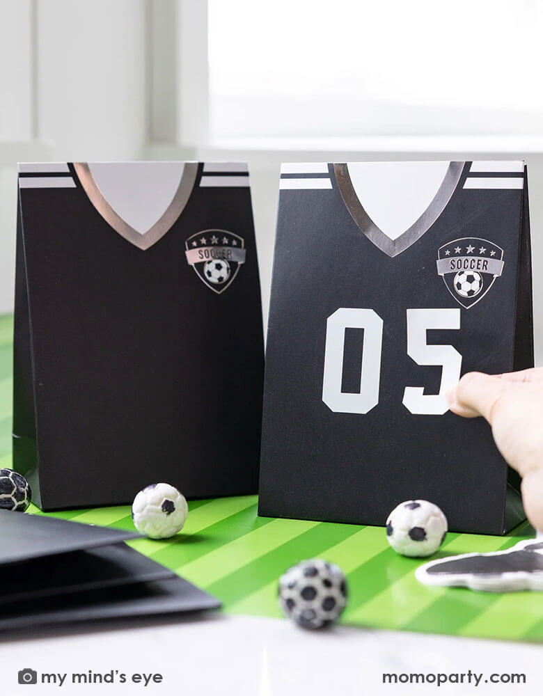 A soccer themed party table features Momo Party's black soccer jersey shaped treat bags with number stickers by My Mind's Eye. On the table there's a green striped soccer table runner and some soccer ball shaped chocolate balls, making it perfect for kid's soccer themed birthday party or championship celebration bash!