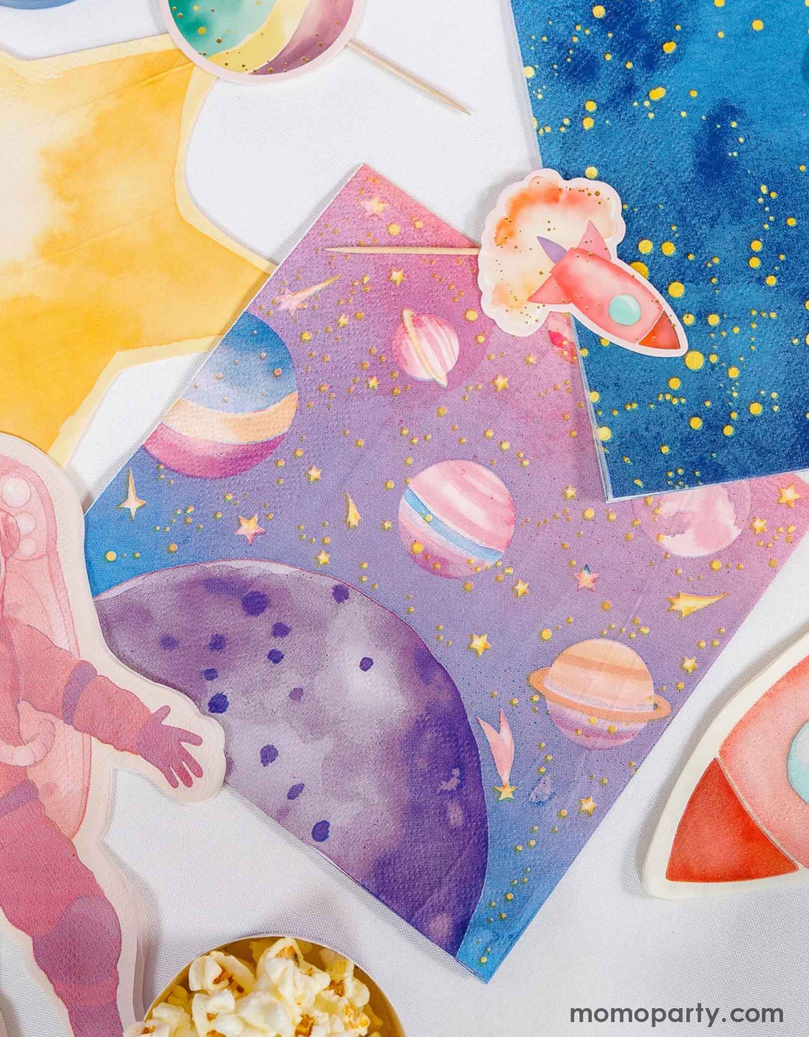 Colorful space-themed party napkins including stars, planets, rocket ship and an astronaut shaped napkin from Momo Party spread out along with some space themed cupcake toppers on a white background.