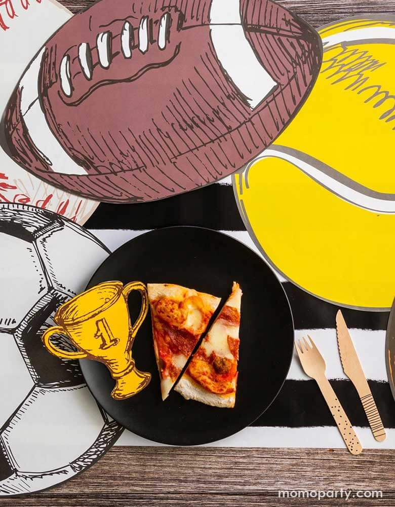 A sports themed party table featuring Momo Party's 7" trophy shaped napkins and a vintage inspired ball shaped placemats including a football placement, a tennis ball shaped placement, a baseball shaped placemat and a soccer ball shaped placemat. On the table there is a plate with a couple of slices of pizzas and some wooden cutleries, all on a black and white striped table runner.
