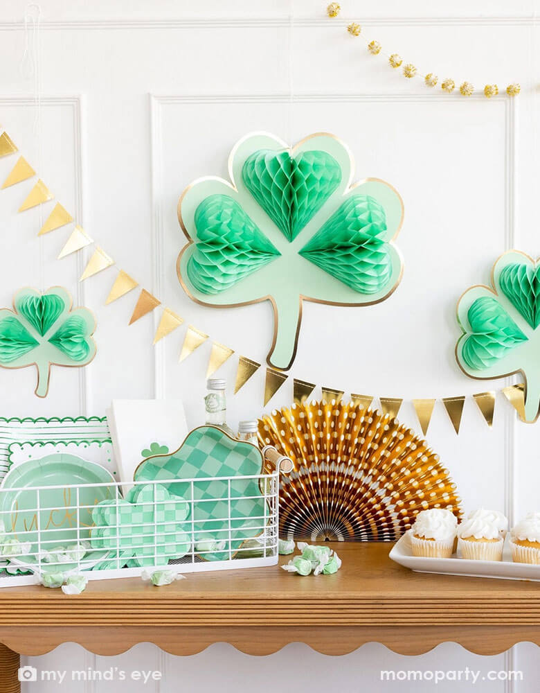 A corner of a room decorated with Momo Party's shamrock honeycomb decorations in various sizes and a gold foil flag banner hung on the wall. On the cabinet counter, there's a gold foil paper party fan, a few cupcakes and a basket filled with Momo Party's St. Patrick's Day party tableware including scalloped paper plates, lucky paper plate set, checkered shamrock shaped napkins and plates next to some green taffy candies, makes this a perfect info for a festive St. Patrick's Day celebration.