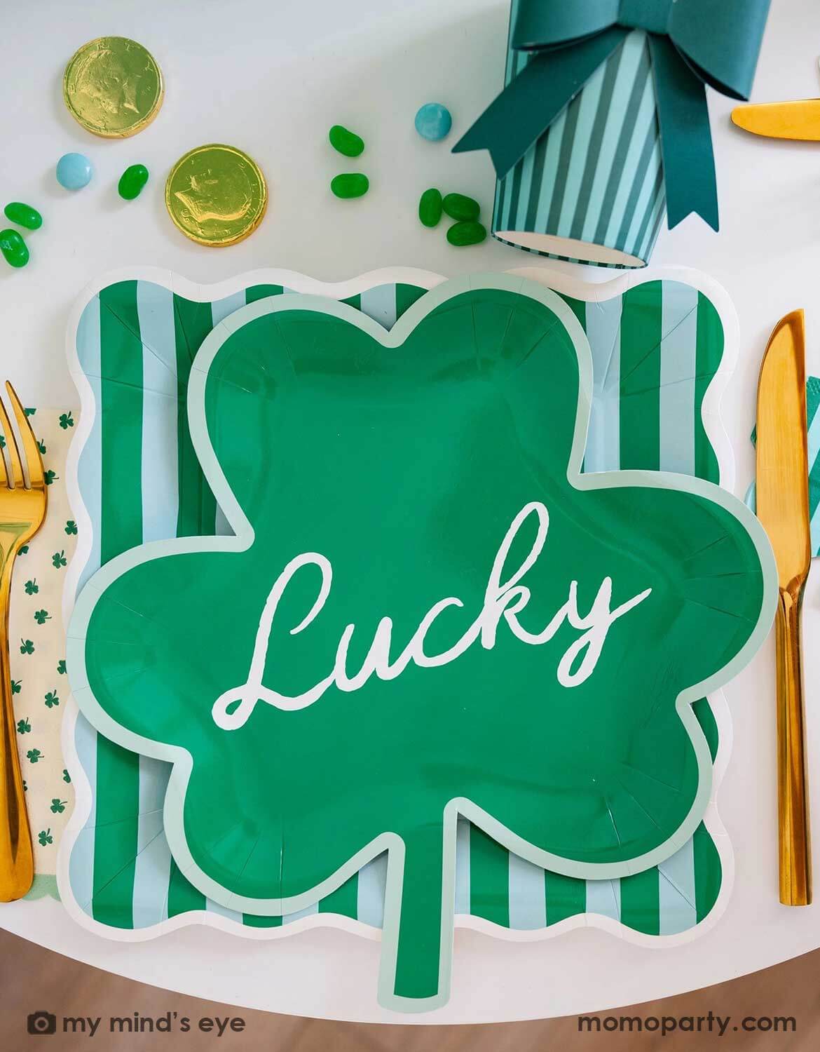 A St. Patrick's Party table setting featuring Momo Party's shamrock-shaped party plate with 'Lucky' text on it on top of a green and light blue striped square plate, with shamrock themed party napkins and party cups around on a festive table setting with gold cutlery, green candies and gold coins. A perfect inspiration for a modern St. Patrick's Day party tablescape.
