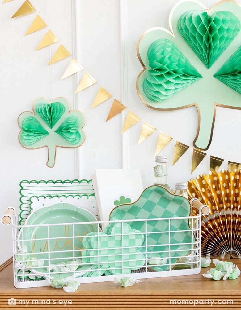 A corner of a house decorated with Momo Party's shamrock honeycomb decorations and a gold foil flag banner hung on the wall. On the cabinet counter, there's a gold foil paper fan and a basket filled with Momo Party's St. Patrick's Day party tableware including scalloped paper plates, lucky paper plate set, checkered shamrock shaped napkins and plates next to some green taffy candies, makes this a perfect info for a festive St. Patrick's Day celebration. 