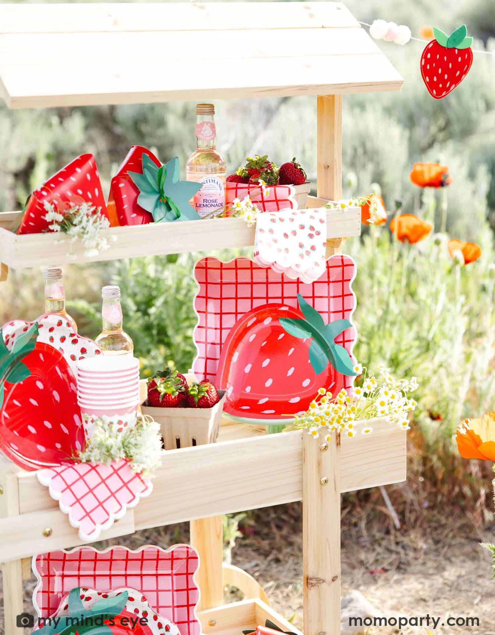 A wooden dessert cart in an outdoor field filled with strawberry-themed tablewares and decorations from Momo Party including strawberry felt banner, strawberry shaped plates, strawberry scattered guest napkins, party cups and treat boxes. Along with bottles, flowers, and strawberries in small baskets, this farmers market-like set up is adorable for kid's "Berry First Birthday" strawberry themed 1st birthday party celebration.