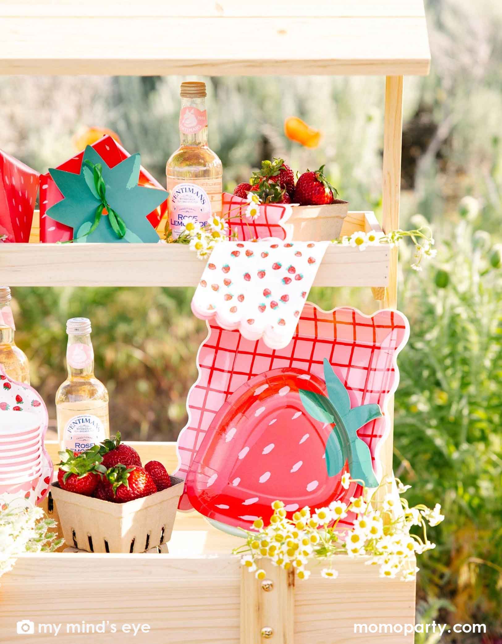 A wooden display cart in an outdoor field filled with strawberry-themed tablewares and decorations from Momo Party including strawberry shaped plates, strawberry scattered guest napkins, party cups and treat boxes. Along with bottles, flowers, and strawberries in small baskets, this farmers market-like set up is adorable for kid's "Berry First Birthday" strawberry themed 1st birthday party celebration.