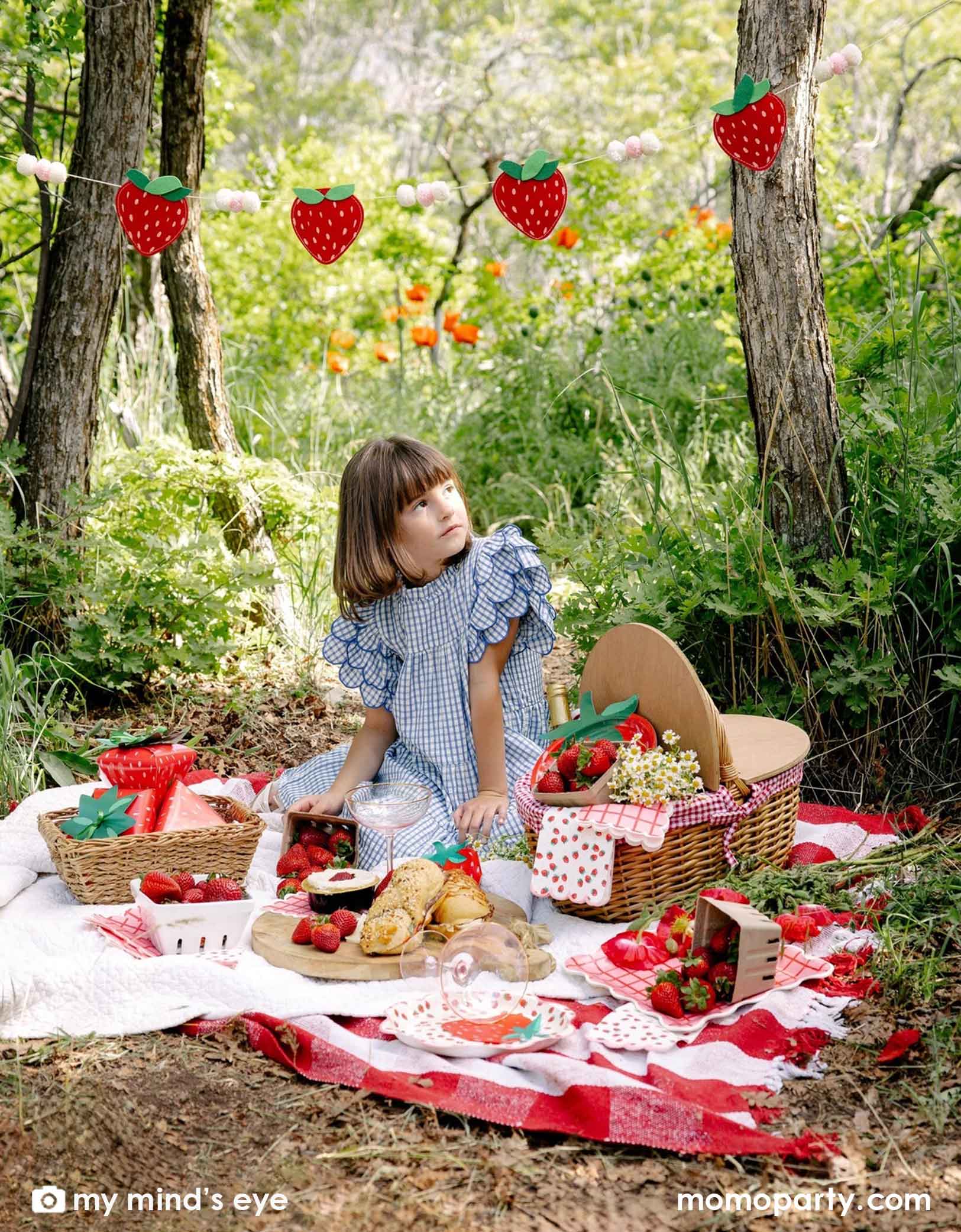 A girl in a blue gingham dress sitting at a picnic blanket in a forest with a decorated area and strawberry decorations and party tablewares from Momo Party including strawberry felt party banner, strawberry plates, napkins, paper cups and strawberry shaped treat boxes. A whimsical and adorable setting for a special celebration. 