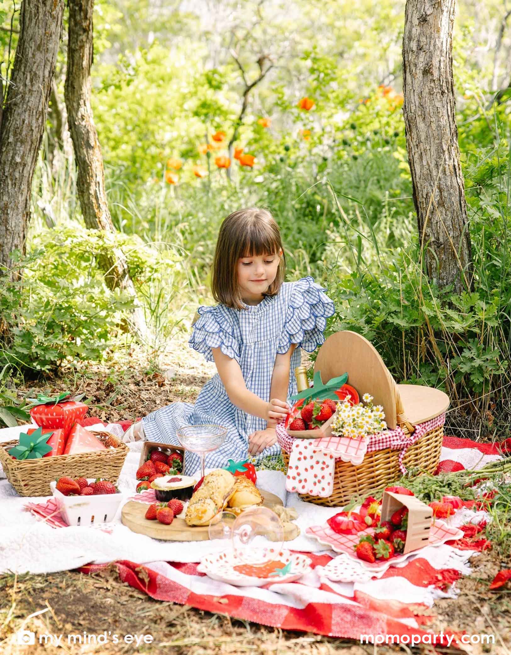 A girl in a blue gingham dress sitting at a picnic blanket with a picnic basket in a forest with a decorated area and strawberry decorations and party tablewares from Momo Party including 10" strawberry plates, napkins, paper cups and strawberry shaped treat boxes. A whimsical and adorable setting for a special celebration. 